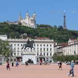 A large city square lined with pavilions, featuring an equestrian statue in the centre, and a church on a tree-covered hill in the background
