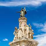 A stone statue showing figures around a pillar, with a metal figure holding a flag on top