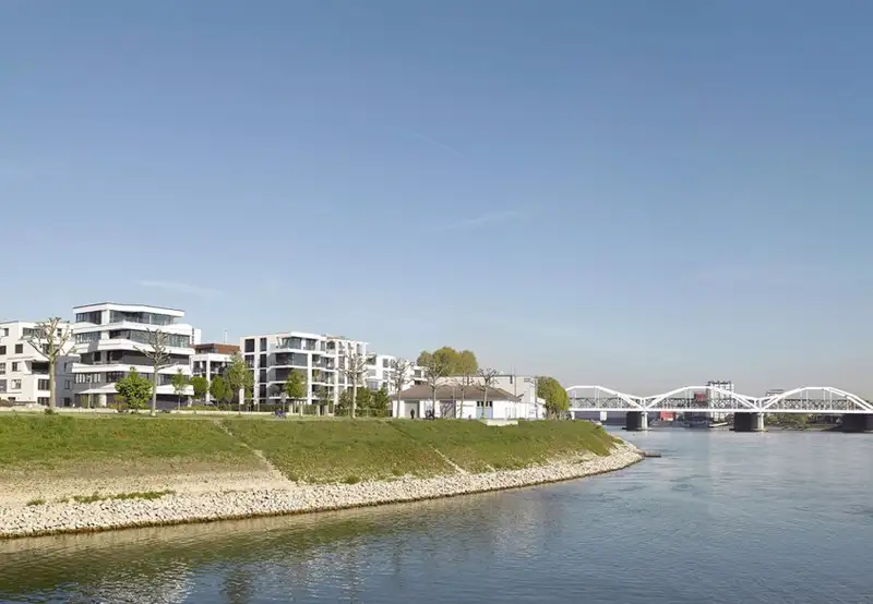 A riverside promenade with a grassy bank and modern mansions in the background